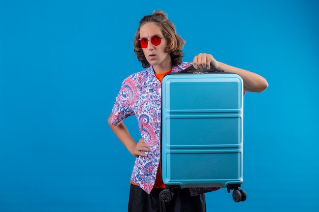 Young handsome guy wearing red sunglasses holding travel suitcase looking at camera surprised and confused standing over blue background