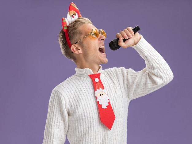 Free Photo young handsome guy wearing santa claus headband and tie with glasses holding microphone singing with closed eyes isolated on purple background with copy space