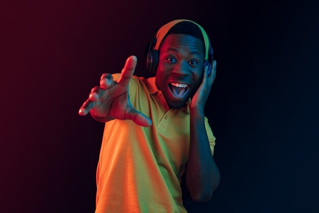 The young handsome happy hipster man listening music with headphones at black studio with neon lights. Disco, night club, hip hop style, positive emotions, face expression, dancing concept