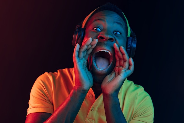 The young handsome happy hipster man listening music with headphones at black studio with neon lights. Disco, night club, hip hop style, positive emotions, face expression, dancing concept