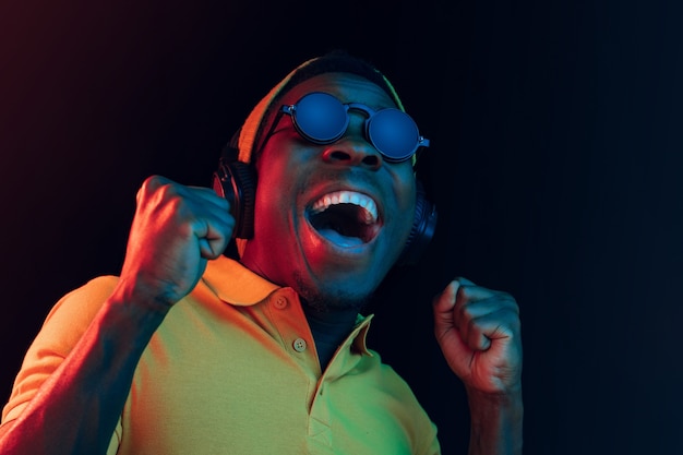 Free photo the young handsome happy hipster man listening music with headphones at black studio with neon lights. disco, night club, hip hop style, positive emotions, face expression, dancing concept