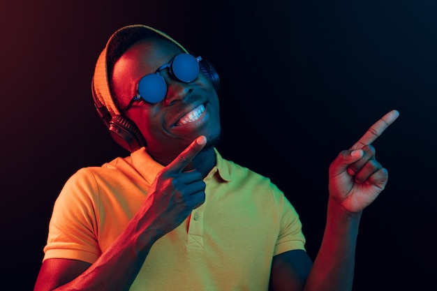 The young handsome happy hipster man listening music with headphones at black studio with neon lights. Disco, night club, hip hop style, positive emotions, face expression, dancing concept