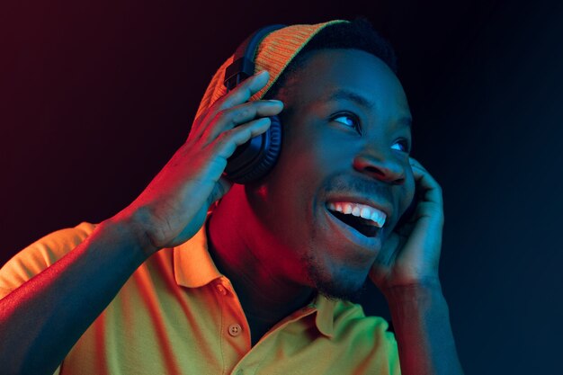 The young handsome happy hipster man listening music with headphones at black studio with neon lights. Disco, night club, hip hop style, positive emotions, face expression, dancing concept