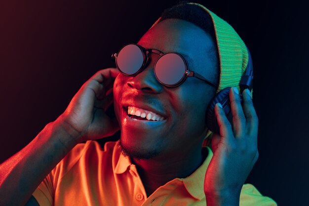 The young handsome happy hipster man listening music with headphones at black studio with neon lights. Disco, night club, hip hop style, positive emotions, face expression, dancing concept