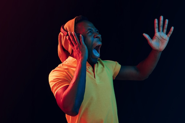 The young handsome happy surprised hipster man listening music with headphones at black studio with neon lights. Disco, night club, hip hop style, positive emotions, face expression, dancing concept