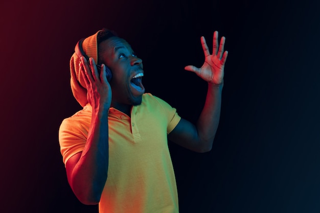 The young handsome happy surprised hipster man listening music with headphones at black studio with neon lights. Disco, night club, hip hop style, positive emotions, face expression, dancing concept