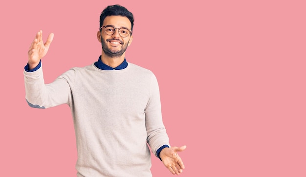 Young handsome hispanic man wearing elegant clothes and glasses looking at the camera smiling with open arms for hug cheerful expression embracing happiness