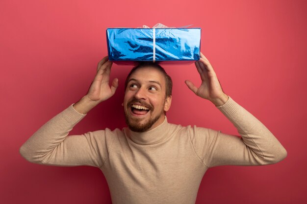 Young handsome man in beige turtleneck holding gist box over his head happy and pleased looking up 