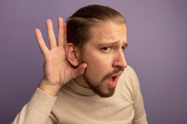 Young handsome man in beige turtleneck holdng hand near his ear trying to listen gossips standing over lilac wall