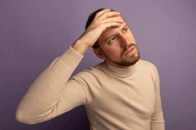 Young handsome man in beige turtleneck looking aside puzzled with hand on his head standing over lilac wall