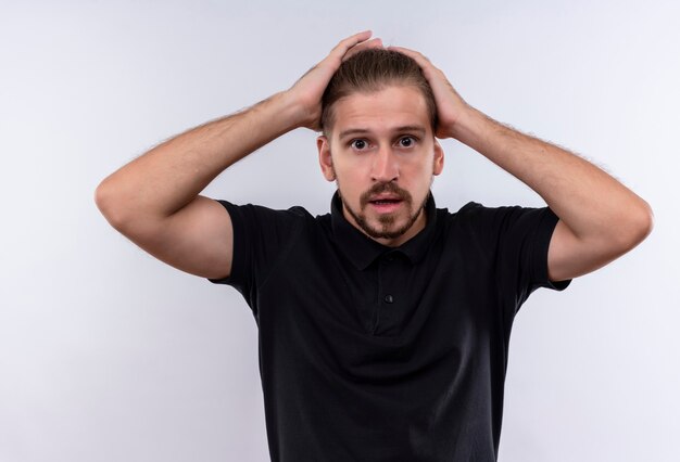 Young handsome man in black polo shirt looking confused and worried touching his head with hands standing over white background