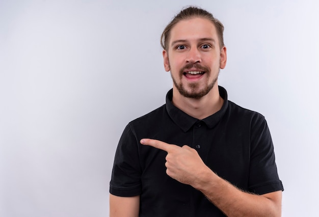 Young handsome man in black polo shirt smiling cheerfully pointing to the side standing over white background