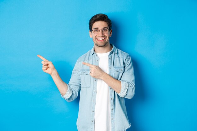 Young handsome man in glasses showing advertisement, smiling and pointing fingers left, making announcement, standing against blue background