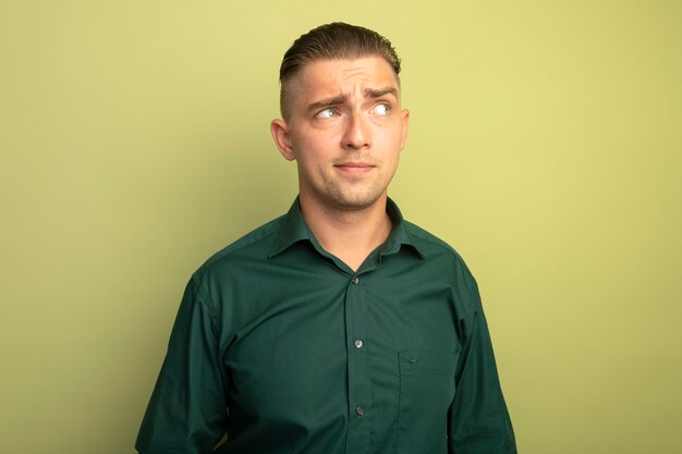 Young handsome man in green shirt looking aside with skeptic expression 