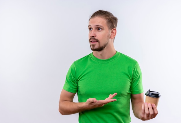 Free photo young handsome man in green t-shirt holding coffee cup looking confused standing over white background