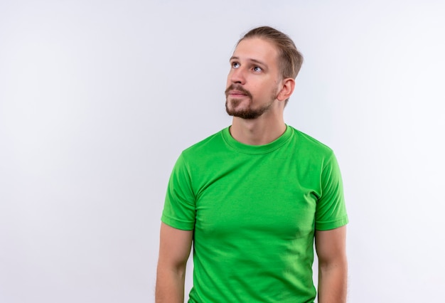 Free photo young handsome man in green t-shirt looking aside with pensive expression on face standing over white background