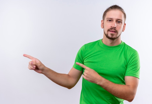 Free photo young handsome man in green t-shirt looking at camera pointing to the side standing over white background