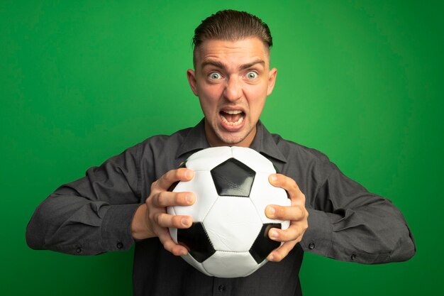 Young handsome man in grey shirt holding soccer ball shouting with aggressive expression 