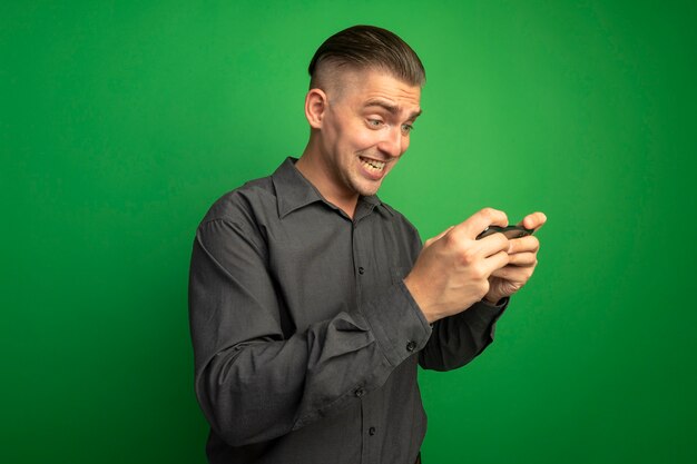 Young handsome man in grey shirt using smartphone happy and excited standing over green wall