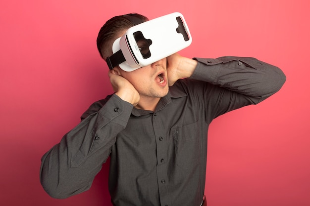 Free photo young handsome man in grey shirt with virtual reality glasses amazed and surprised standing over pink wall