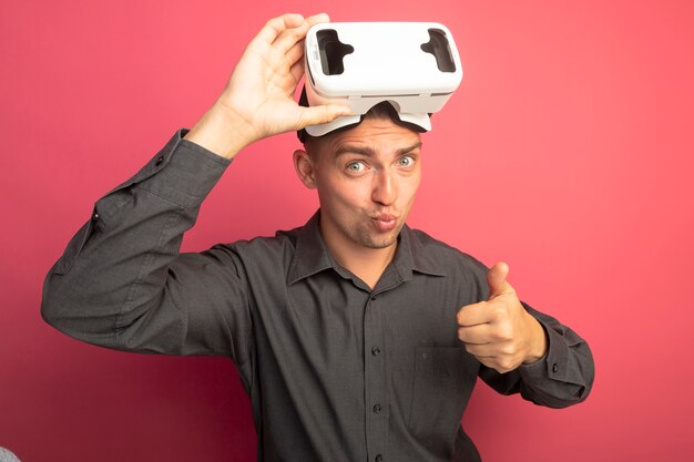 Young handsome man in grey shirt with virtual reality glasses looking confident showing thumbs up standing over pink wall