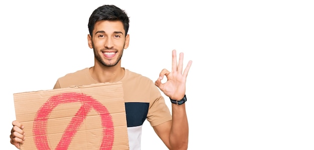 Young handsome man holding prohibited warning banner doing ok sign with fingers, smiling friendly gesturing excellent symbol