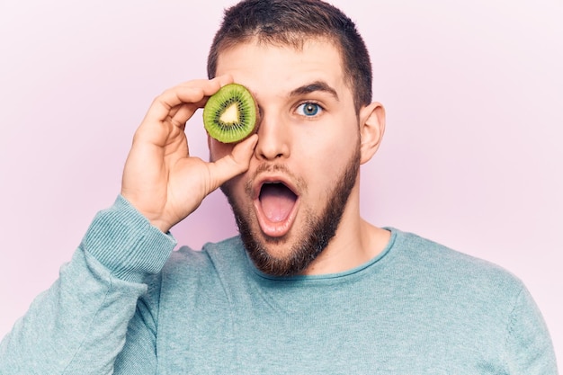 Free photo young handsome man holding slice of kiwi over eye scared and amazed with open mouth for surprise disbelief face