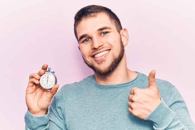 Free photo young handsome man holding stopwatch smiling happy and positive thumb up doing excellent and approval sign