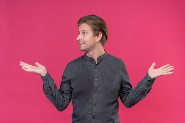 Free photo young handsome man looking confused spreading arms smiling having no answer standing over pink wall