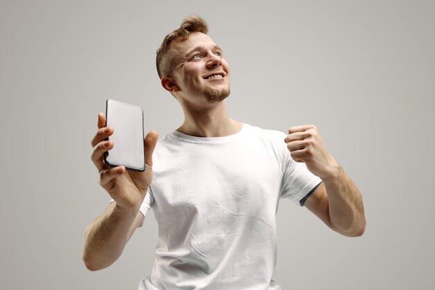 Young handsome man showing smartphone screen over gray background with a surprise face. Human emotions, facial expression concept