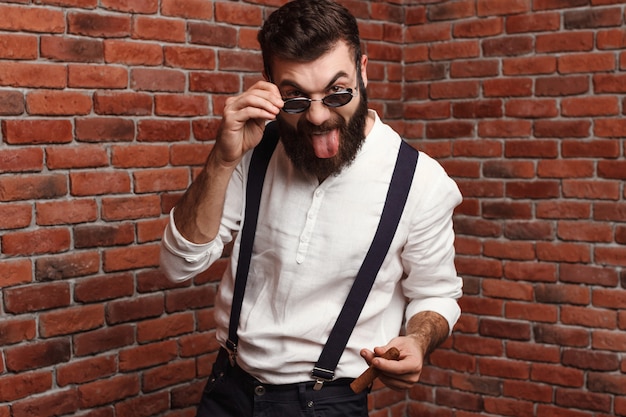 Free Photo young handsome man showing tongue holding cigar on brick wall.