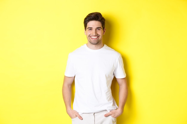 Young handsome man smiling at camera, holding hands in pockets, standing against yellow background.