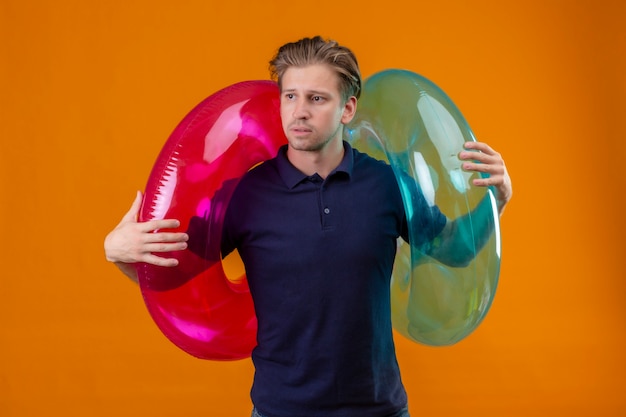 Free Photo young handsome man standing with inflatable rings looking aside with sad expression on face over orange background