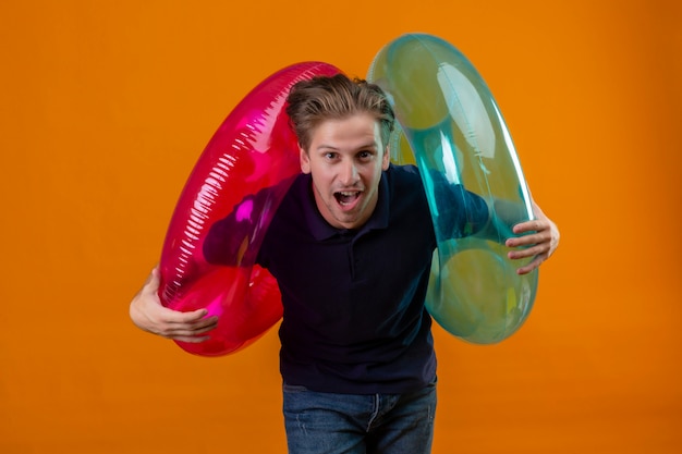 Free photo young handsome man standing with inflatable rings surprised and happy with wide open mouth over orange background