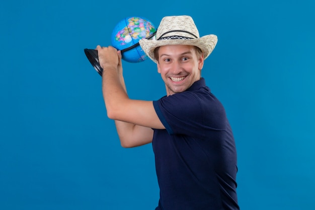 Young handsome man in summer hat holding globe threatening to hit with globe joking and smiling standing over blue background