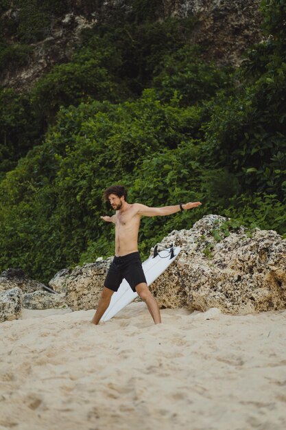 A young handsome man surfer on the ocean shore is doing a warm-up before surfing. exercises before sports, stretching before surfing.