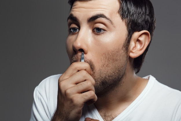 Young handsome man tweezing hair in nose over grey wall