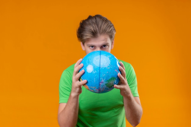 Free Photo young handsome man wearing green t-shirt holding globe hiding behind it with serious face standing over orange wall