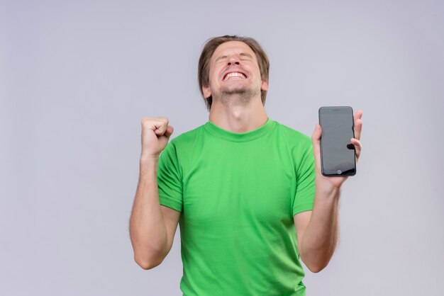 Young handsome man wearing green t-shirt holding mobile phone crazy happy clenching fist rejoicing his success standing over white wall