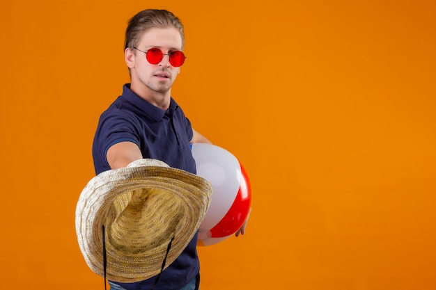 Free photo young handsome man wearing red sunglasses holding inflatable ball and stretching out straw hats with confident expression standing over orange background