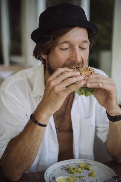 Young handsome man in a white shirt open, having breakfast in a cafe with a vegetarian burger, drinking coffee, lifestyle in a tropical island, life in Bali.