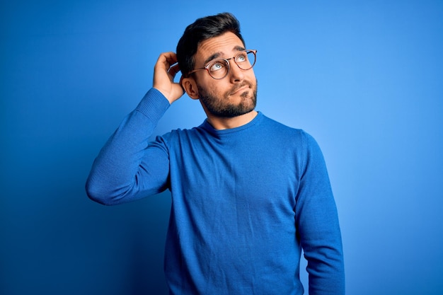 Free Photo young handsome man with beard wearing casual sweater and glasses over blue background confuse and wondering about question uncertain with doubt thinking with hand on head pensive concept