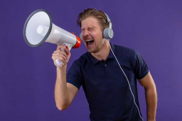 Young handsome man with headphones shouting through megaphone standing over purple background