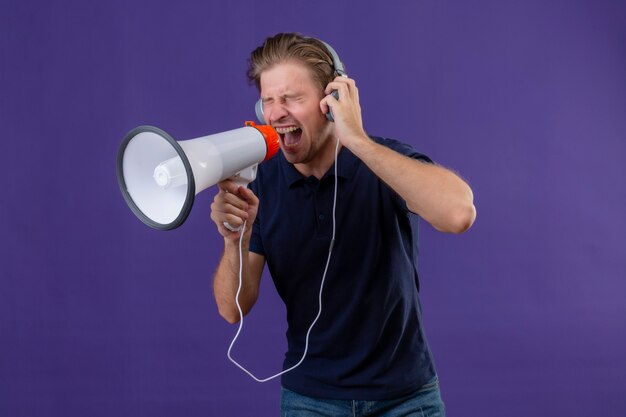 Young handsome man with headphones shouting through megaphone standing over purple background