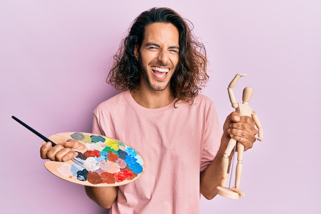 Free photo young handsome man with long hair holding painter palette and art manikin smiling and laughing hard out loud because funny crazy joke.