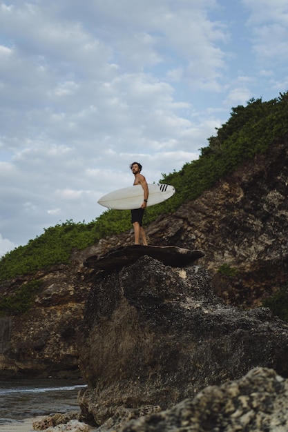 Young handsome man with a surfboard on a rock near the ocean.