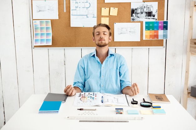 Free photo young handsome pensive relaxed businessman meditating sitting at table. white modern office interior.