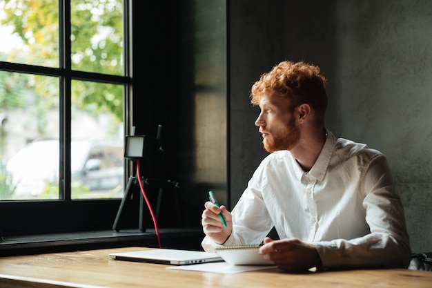 Young handsome readhead bearded man sitting on the workplace at home, looking at big window