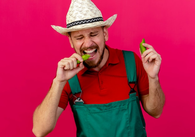 Young handsome slavic gardener in uniform and hat holding and biting pepper with closed eyes 