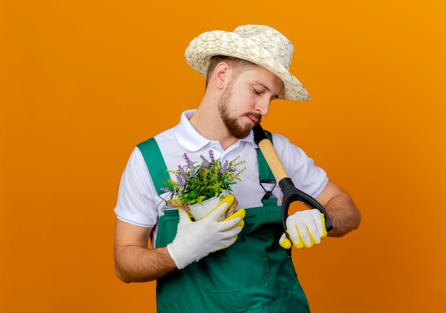 Young handsome slavic gardener in uniform wearing hat and gardening gloves holding spade and flowerpot looking at spade isolated